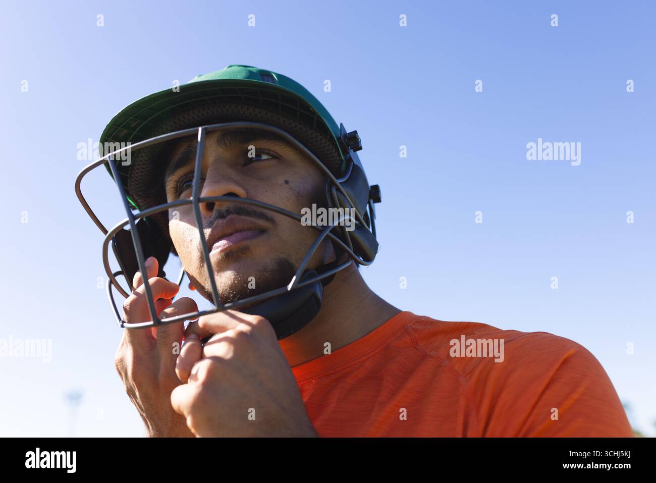 Indischer Mann, der den grünen Cricket-Helmgurt im orangefarbenen Sporthemd auf dem Sportplatz anzieht Stockfoto
