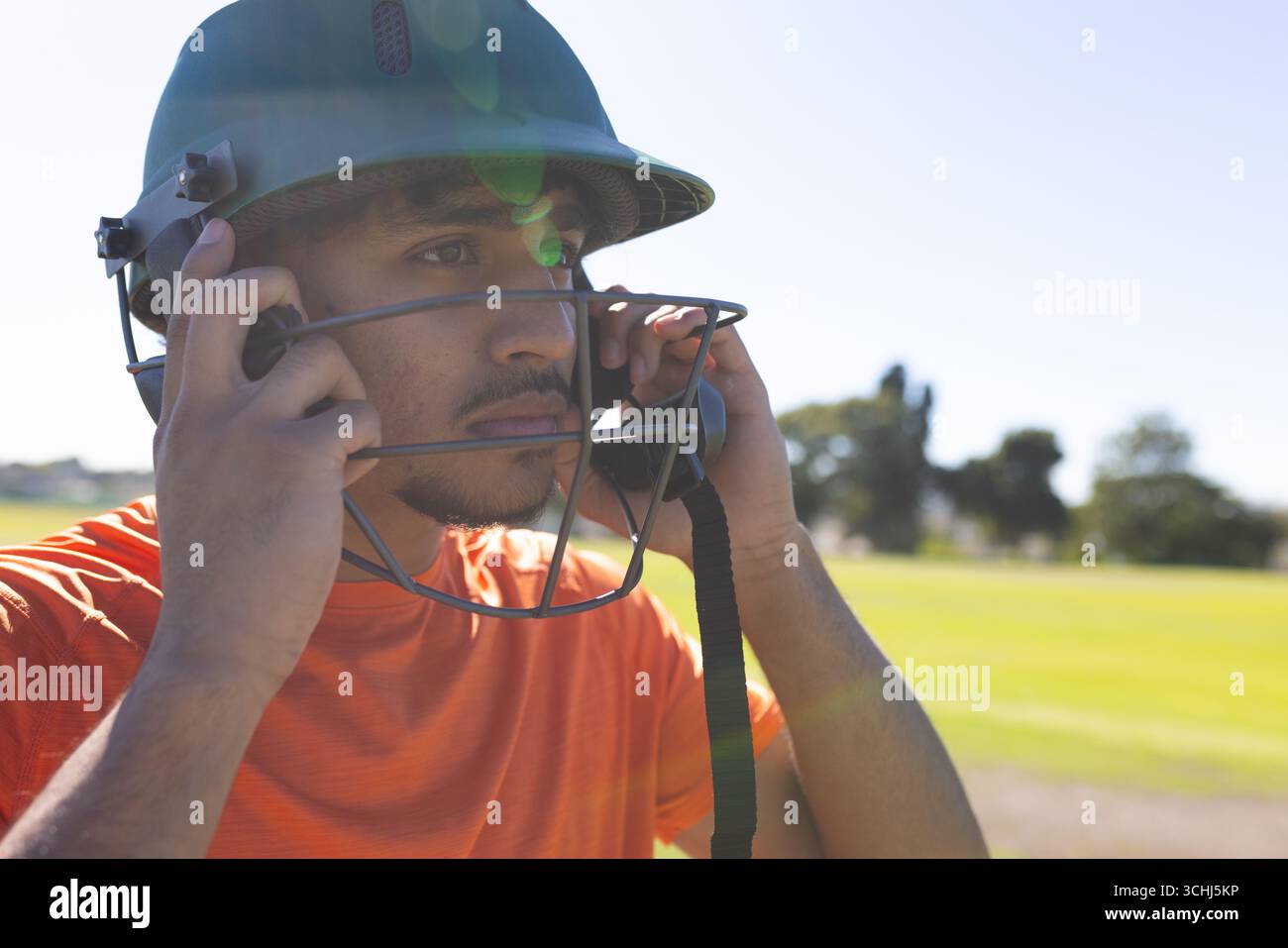 Indischer Mann im orangefarbenen Sportrikot auf dem Cricketfeld, der den Kinnriemen des Helms hält Stockfoto