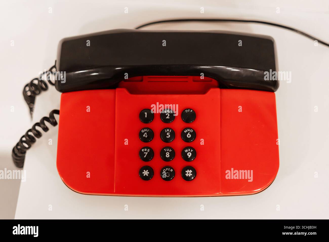 Ein klassisches rotes Drehtelefon liegt auf einem weißen Tisch und zeigt sein markantes schwarzes Mobilteil und sein auffälliges Design. Er verleiht dem einen Retro-Touch Stockfoto