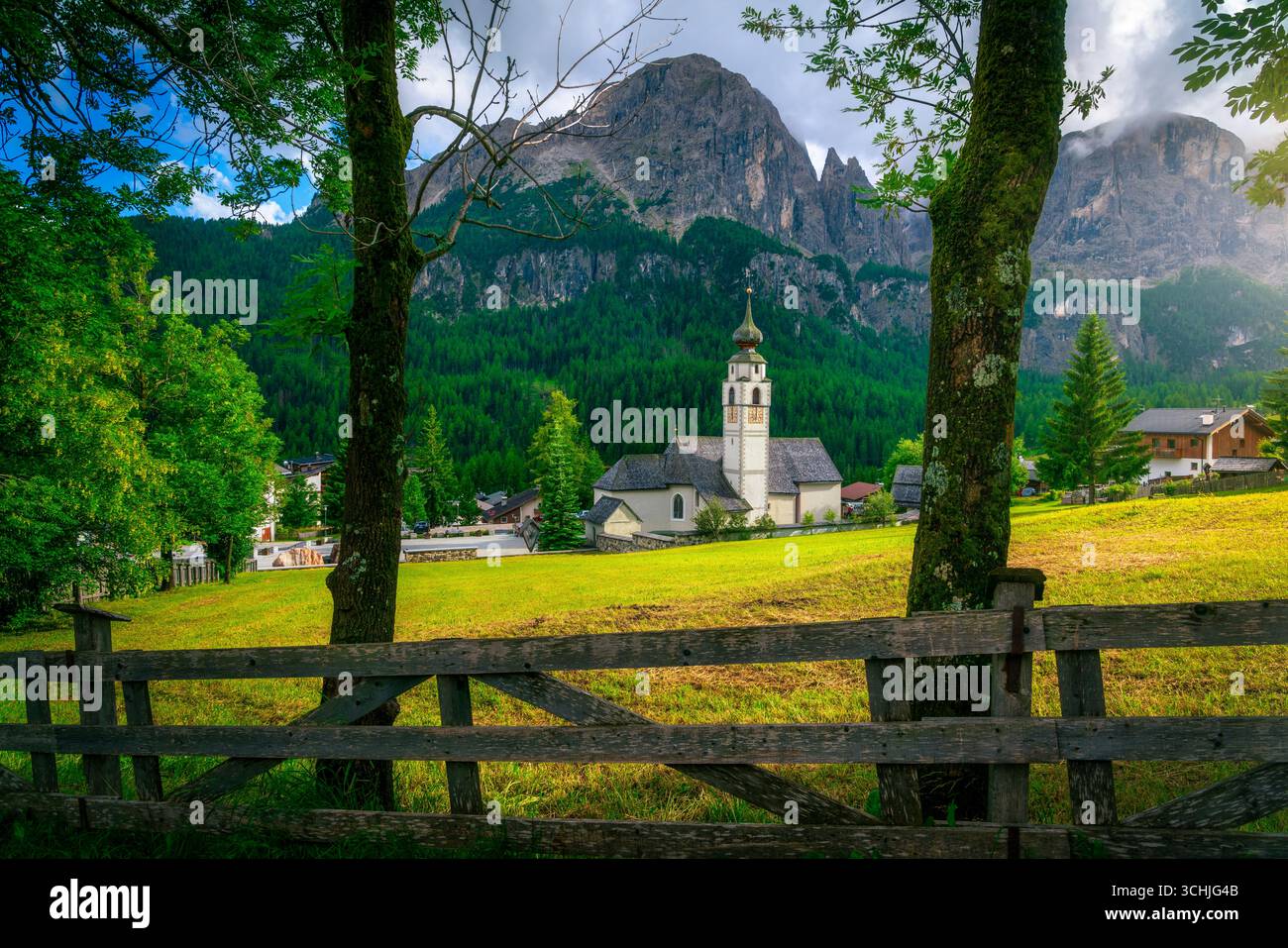 Malerische Bergkirche im Dorf Colfosco, eingerahmt von Holzzaun und Bäumen mit hoch aufragenden Dolomiten Kalksteinklippen und grünen Wiesen bei Sonnenuntergang. S Stockfoto