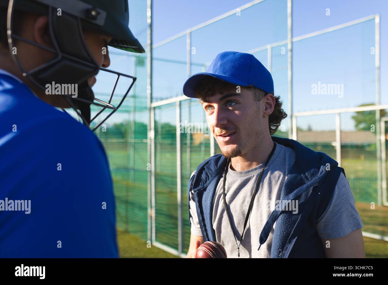 Männlicher Trainer mit Cricketball und leitender männlicher Spieler, der im Übungsnetz auf dem Rasenfeld zuhört Stockfoto