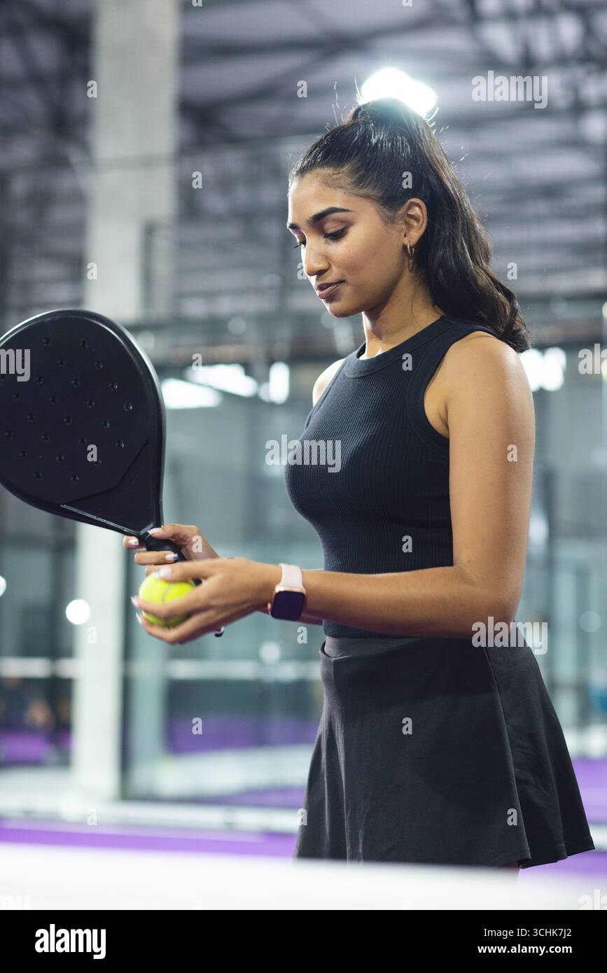 Indische Frau packt Padel-Schläger und konzentriert sich auf gelben Ball in Smartwatch auf Padel Court Stockfoto