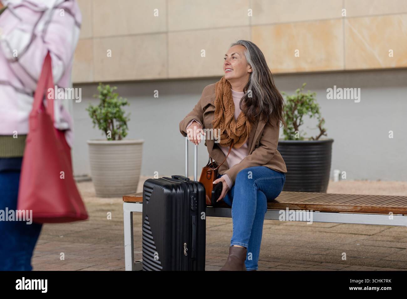 Reife Erwachsene Frau, die auf einer Bank vor dem Gebäude sitzt und einen schwarzen Koffer hält, mit einer Handtasche Stockfoto