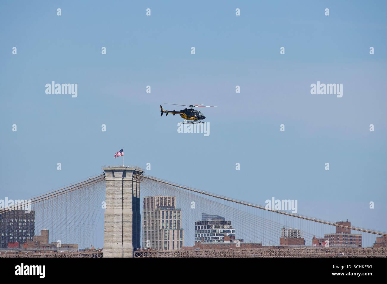 Schwarz-gelber Hubschrauber, der an einem sonnigen Sommertag tief an der Brooklyn Bridge vorbeifliegt und mit amerikanischer Flagge oben über den Hafen von New York fährt Stockfoto