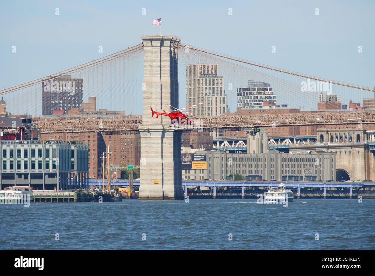 Roter Hubschrauber fliegt tief an der Brooklyn Bridge vorbei mit amerikanischer Flagge oben, über den New Yorker Hafen an einem sonnigen Sommertag mit blauem Himmel, Kopierraum Stockfoto