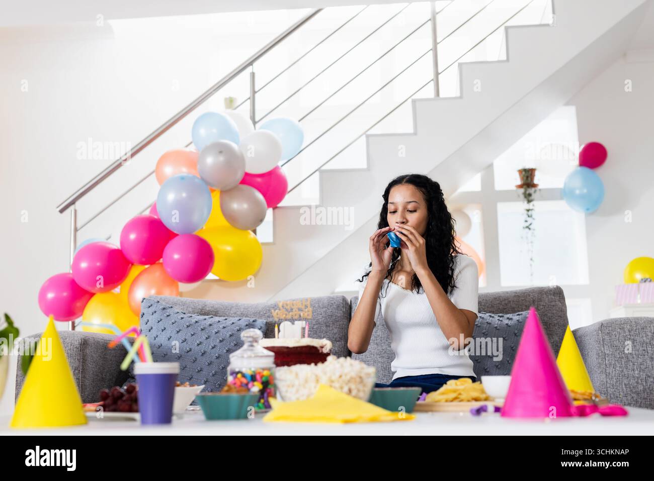 Zu Hause feiert die Frau das Partyhorn, umgeben von bunten Ballons Stockfoto