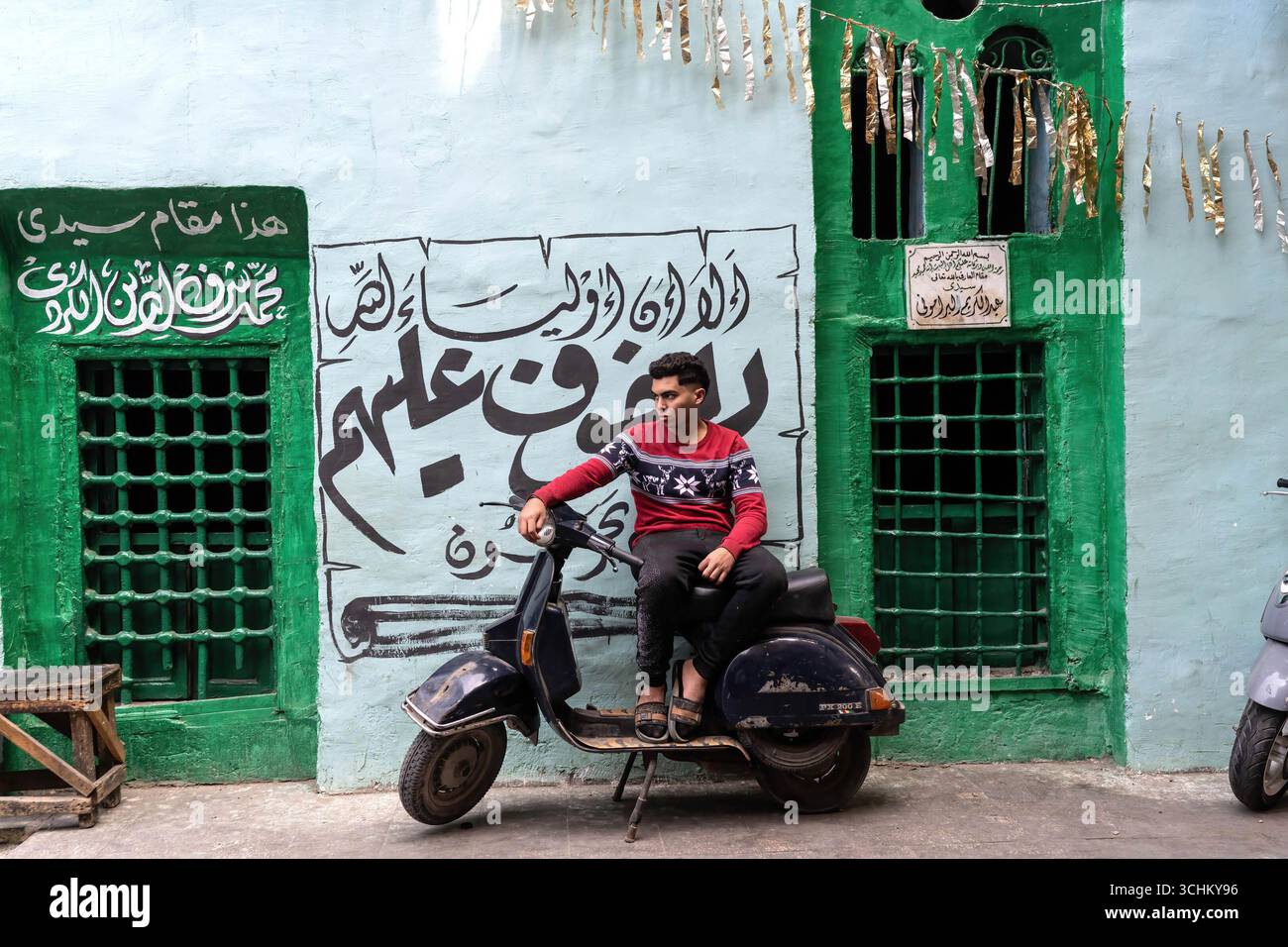 Ein junger ägyptischer Mann sitzt auf einem Motorroller vor einem hell bemalten Grab mit arabischen Inschriften im islamischen Viertel in der Nähe von Midan Hussein und dem Khan el Khalili Basar in Kairo. Stockfoto