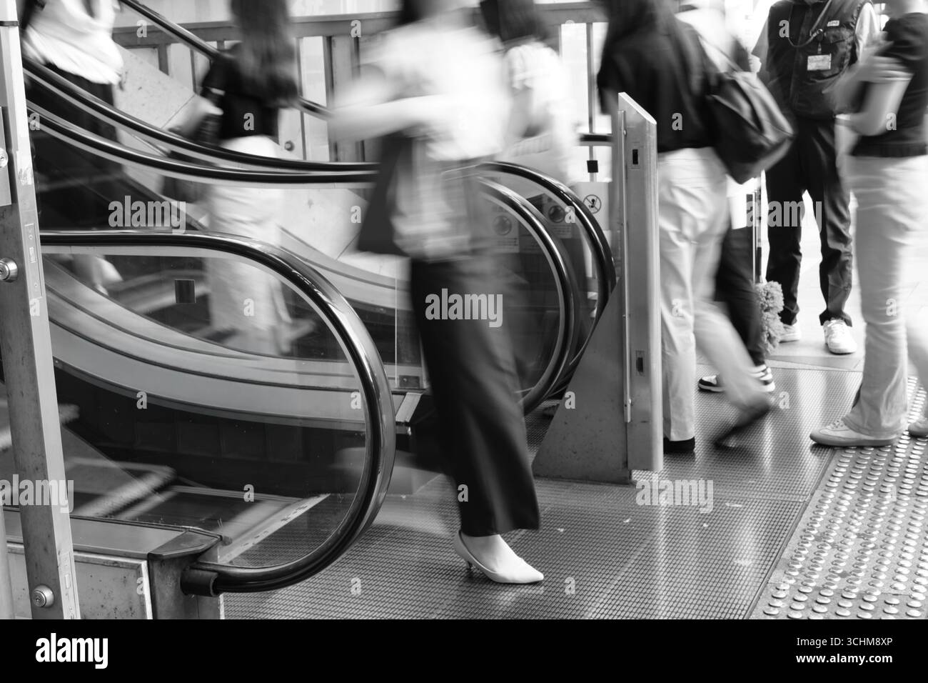Frauen in Bewegung verlassen eine Rolltreppe in einer schwarz-weißen Stadt, die urbanen Rhythmus, Alltag und flüchtige Konzentration in einer temporeichen Umgebung zeigt Stockfoto