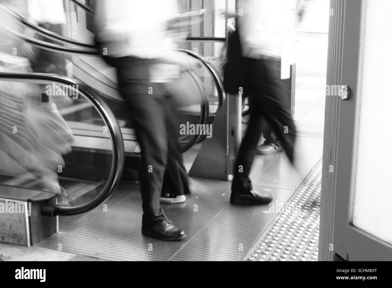 Verschwommene Geschäftsleute verlassen eine Rolltreppe in einem stark frequentierten städtischen Raum und spiegeln so den Rhythmus des beruflichen Alltags wider. Stockfoto