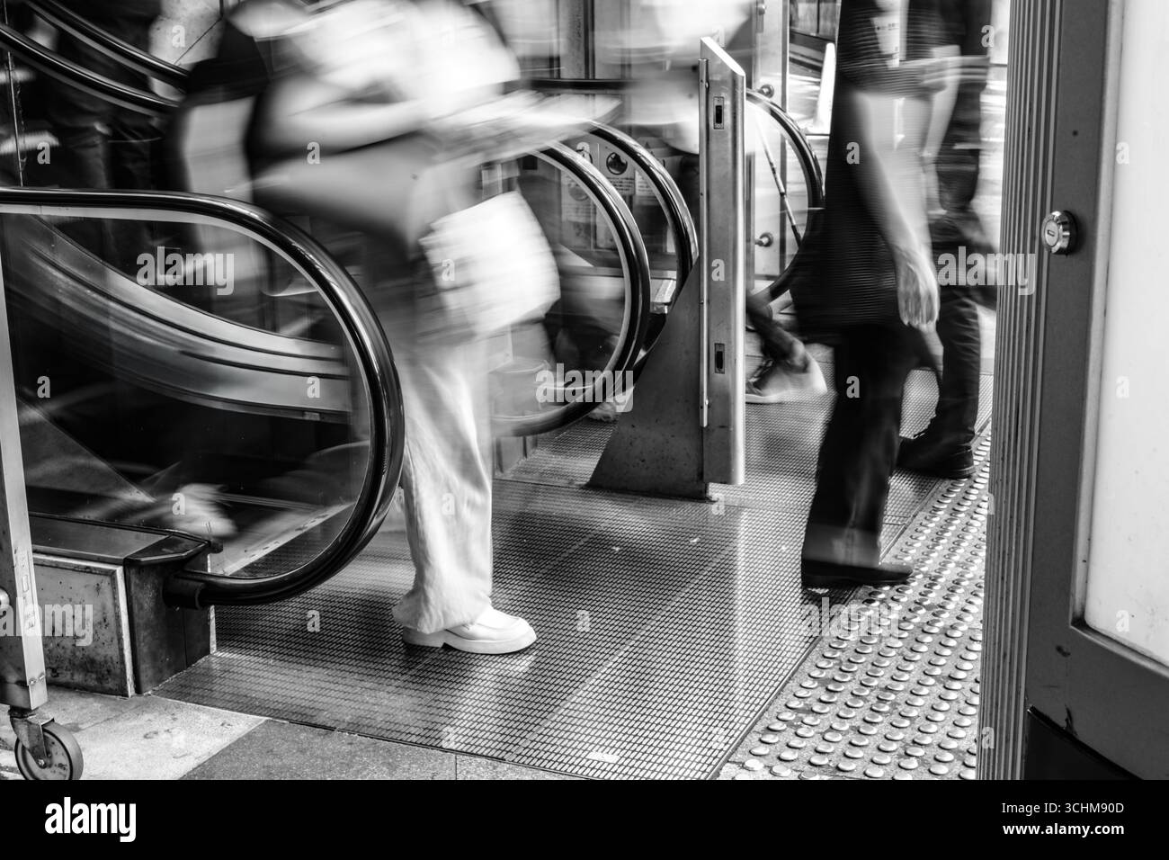 Bewegungsunscharfe Frau verlässt die Rolltreppe in einer geschäftigen Stadtszene und fängt das flüchtige Tempo des urbanen Lebens ein. Schwarzweißtöne Stockfoto