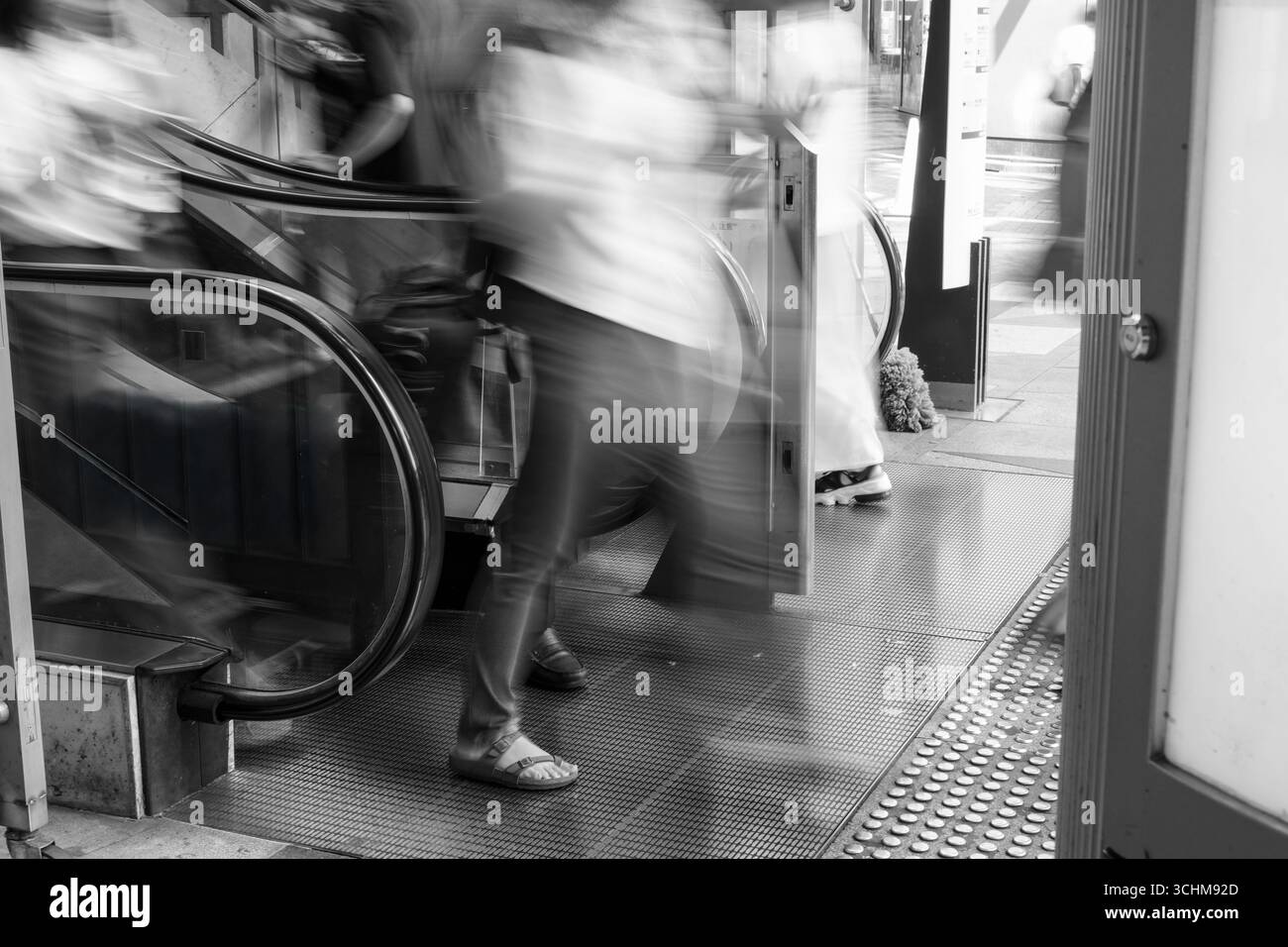 Bewegungsunscharfe Frau verlässt die Rolltreppe in einer geschäftigen Stadtszene und fängt das flüchtige Tempo des urbanen Lebens ein. Schwarzweißtöne betonen die Geschwindigkeit Stockfoto
