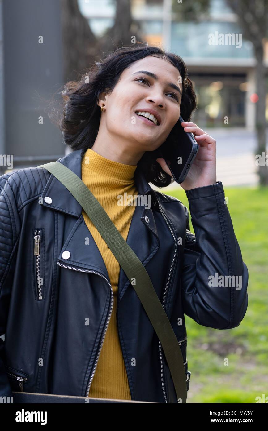 Asiatische Frau lächelt, während sie ihr Smartphone in Lederjacke im Stadtpark hält Stockfoto