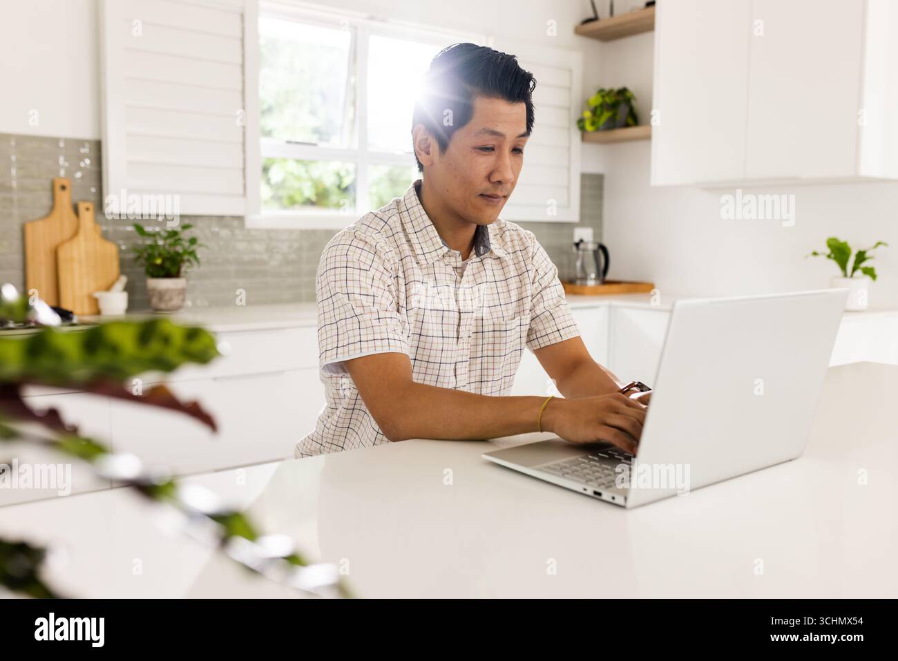 Asiatischer Mann mit Laptop in heller Küche, konzentriert sich auf die Arbeit, zu Hause Stockfoto