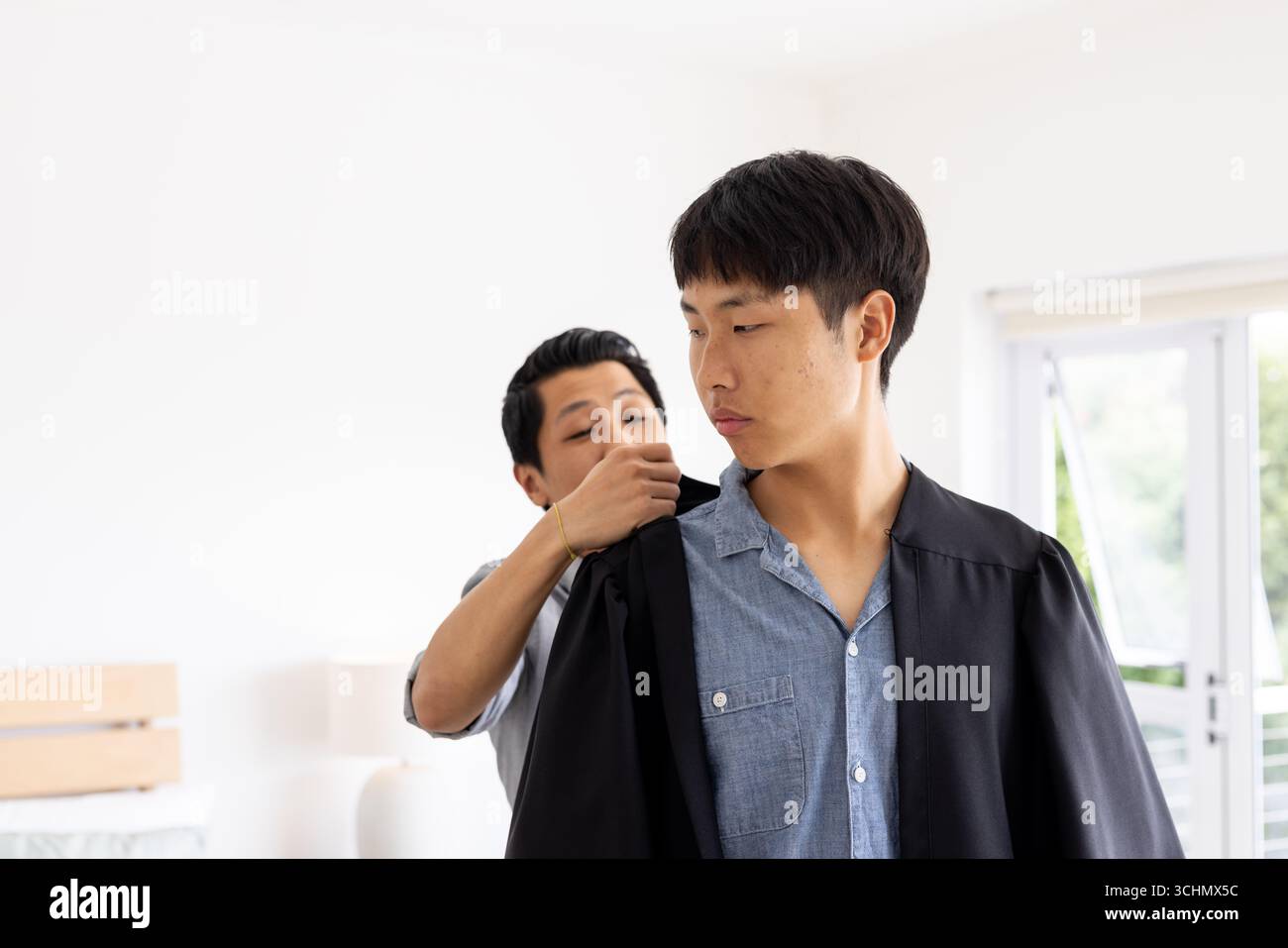 Vater hilft dem Sohn, zu Hause ein Abschlusskleid anzuziehen, beide sehen stolz aus, Kopierraum Stockfoto