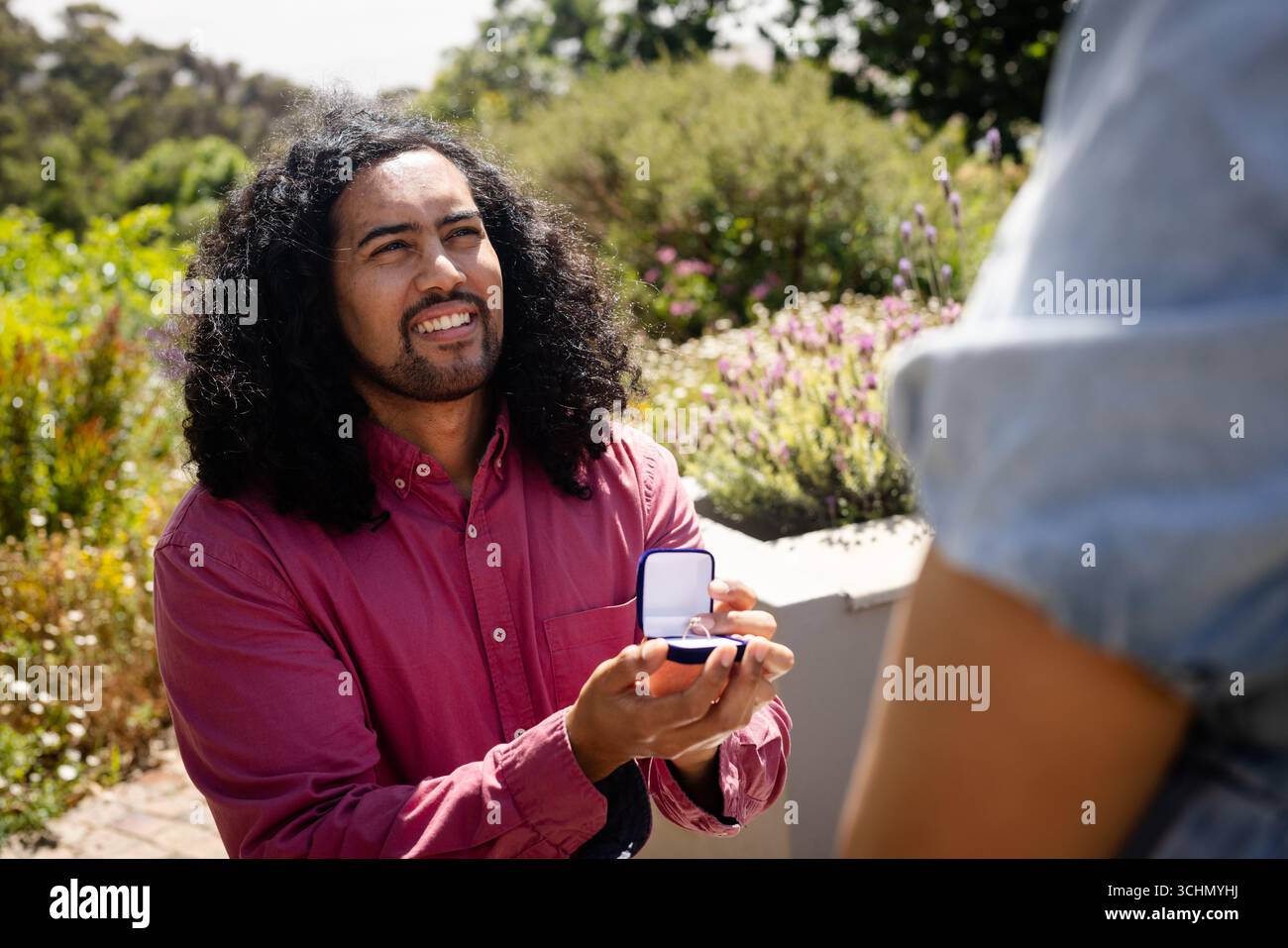 Ein Mann, der mit einer Ringbox im Garten einen Antrag macht, lächelt und hoffnungsvoll aussieht Stockfoto