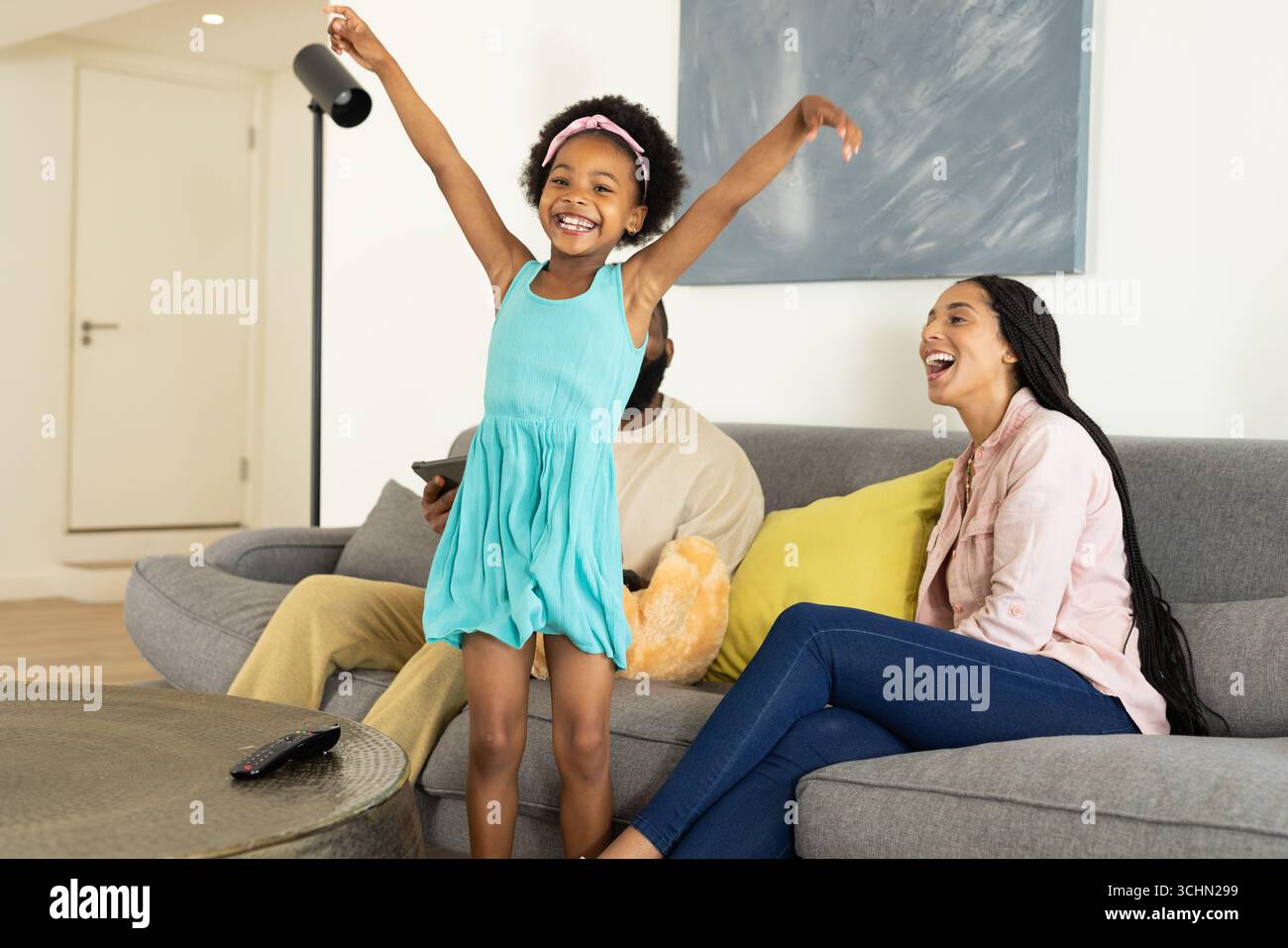 Fröhliches Mädchen in blauem Kleid, das zu Hause tanzt und die Familie lächelt auf der Couch Stockfoto