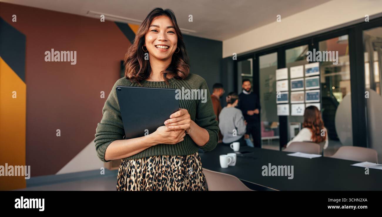 Eine lächelnde Frau steht vor einem kreativen Büro, während ihr Team im Hintergrund zusammenarbeitet. Hebt Führung, Teamarbeit und Dynamik hervor Stockfoto