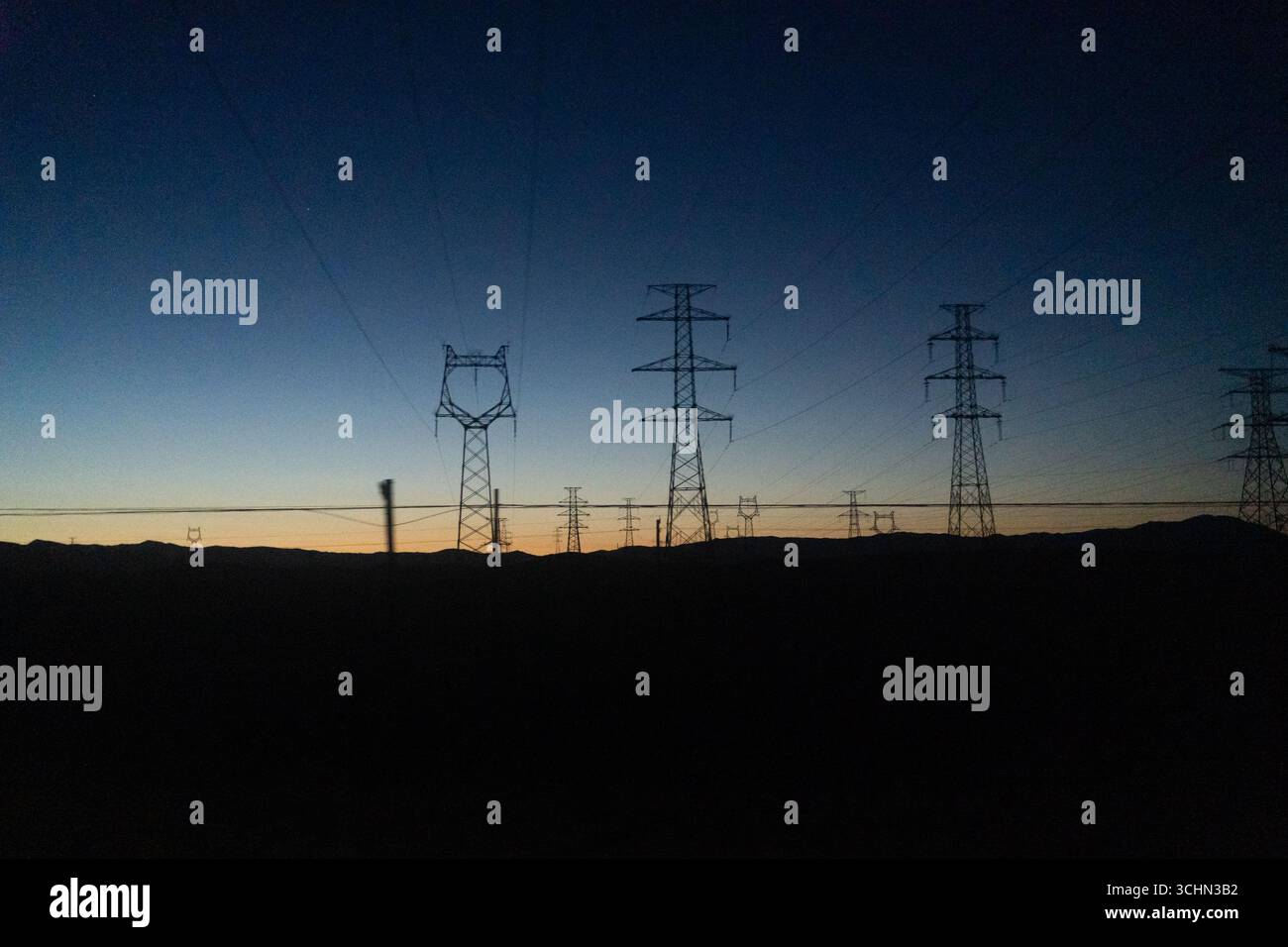 Silhouetten von Hochspannungs-Stromübertragungstürmen und -Linien vor einem farbenfrohen Sonnenuntergang über dunklen Hügeln. Stockfoto