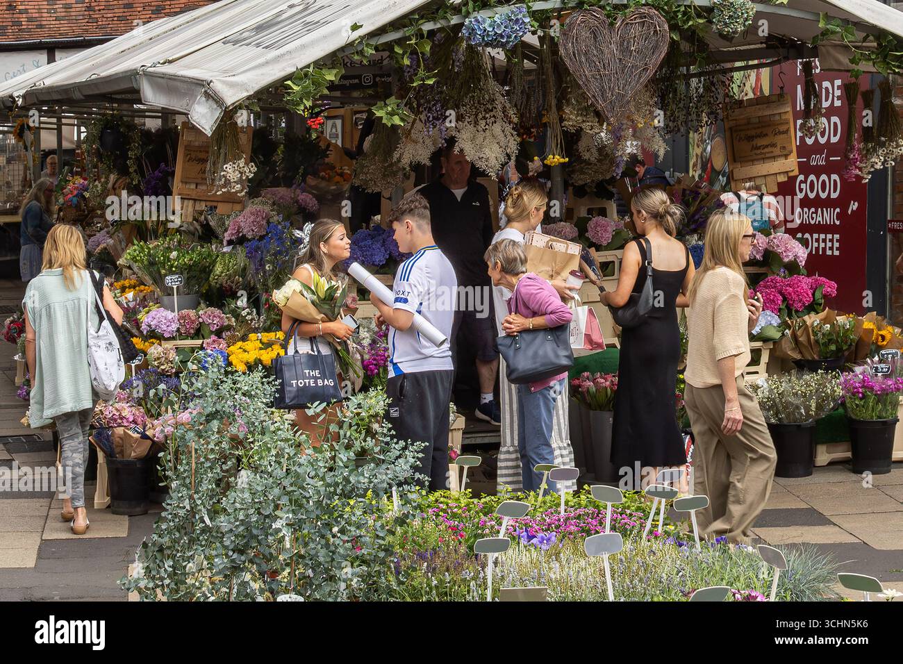 Ein belebter Blumenstand auf einem Gemeinschaftsmarkt mit lebhaften Blüten, handgefertigter Einrichtung und Shopping-Besuchern, die in einer lebhaften und einladenden Atmosphäre stöbern. Stockfoto