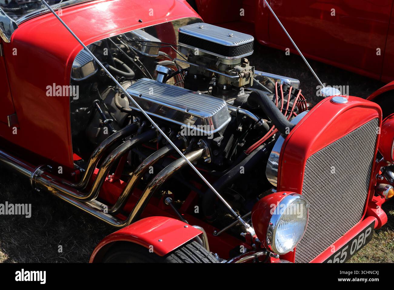 Diese Abbildung zeigt den belichteten V8-Motor eines roten Hot Rod aus der Nähe und zeigt die polierten Chrom-Ansaugkrümmer und -Schneidwerke. Das Auto mit seinem klassischen Kühlergrill und den Scheinwerfern wird auf der Stokes Bay Car Rallye gezeigt. Die Rohleistung des Motors wird durch die sichtbaren Komponenten hervorgehoben. Illustrativ, Editorial: Die Stokes Bay Car Rally in Gosport, Hampshire, England. 25. August 2025. Stockfoto