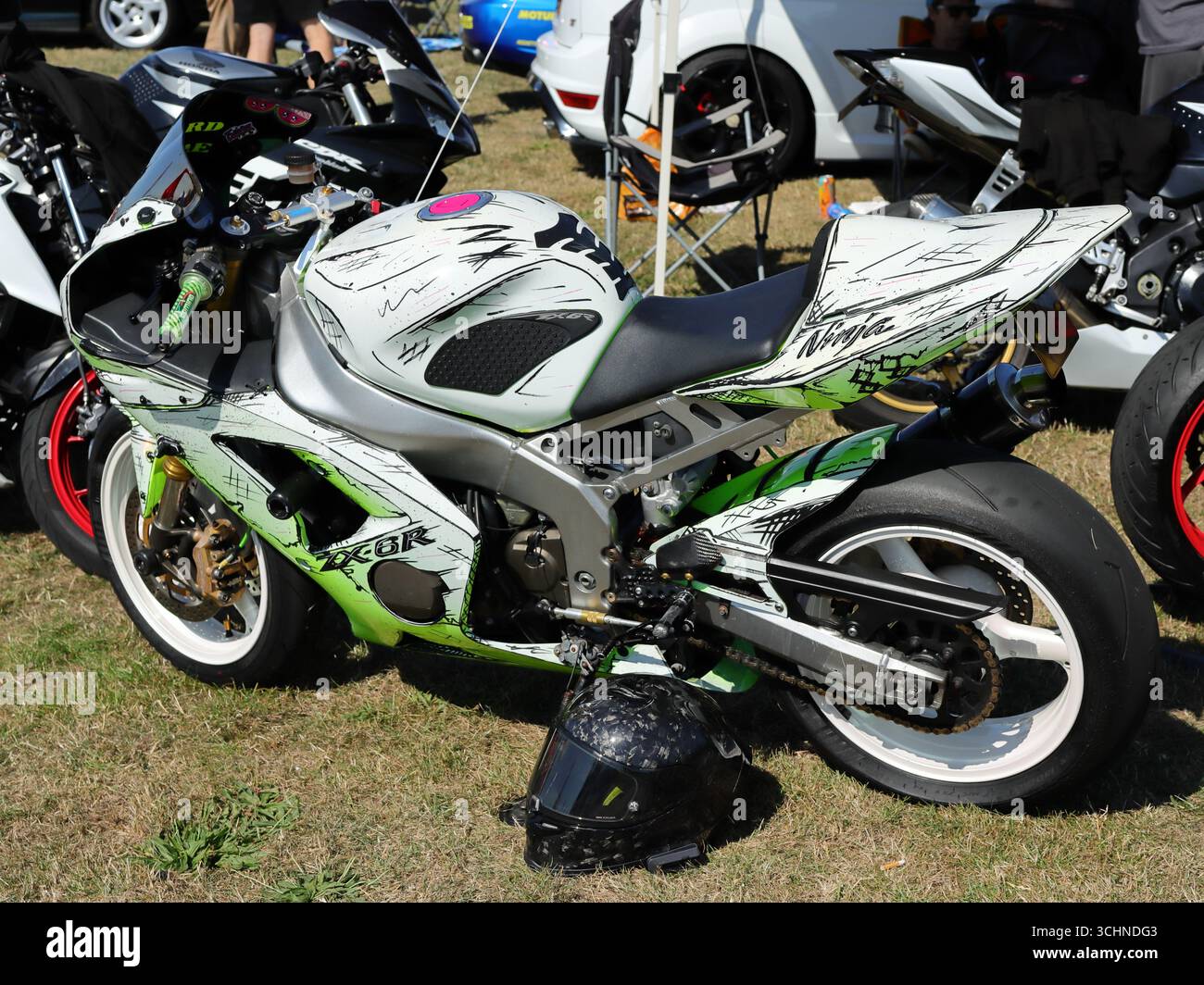 Dieses Bild zeigt ein Kawasaki Ninja Sportmotorrad mit einem benutzerdefinierten grün-weißen Grafikdesign. Neben dem Fahrrad befindet sich ein Helm auf dem Boden und ein großer Hinterreifen. Andere Motorräder und Autos sind im Hintergrund sichtbar und zeigen die Rallye-Einstellung. Illustrativ, Editorial: Die Stokes Bay Car Rally in Gosport, Hampshire, England. 25. August 2025. Stockfoto