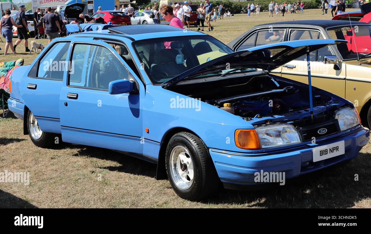 Dieses Bild zeigt eine blaue Ford Sierra Cosworth Limousine mit geöffneter Motorhaube bei der Stokes Bay Car Rally. Das Fahrzeug verfügt über spezielle Leichtmetallfelgen, ein Schiebedach und die Fahrertür ist offen. Ein anderer Oldtimer ist teilweise im Hintergrund zu sehen. Illustrativ, Editorial: Die Stokes Bay Car Rally in Gosport, Hampshire, England. 25. August 2025. Stockfoto