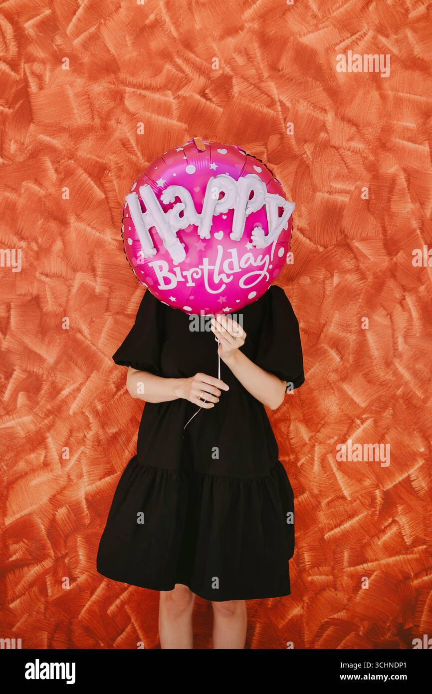 Eine Frau mittleren Alters hält einen übergroßen rosa Heliumballon mit den Worten Happy Birthday auf ihrem Gesicht, was einen verspielten und unbeschwerten Moment schafft Stockfoto