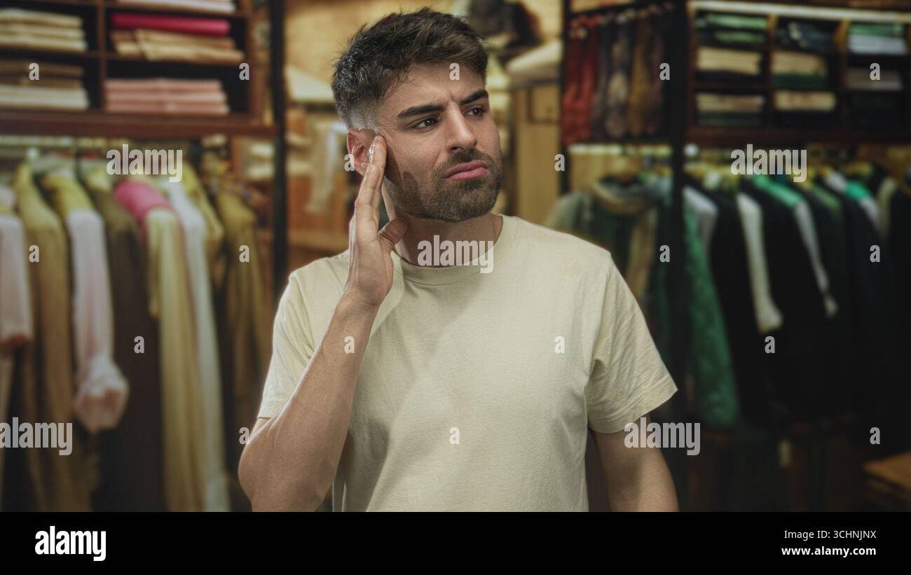 Ein Mann mit weißem T-Shirt berührt die Wangengeste beim Bau eines Bekleidungsgeschäfts mit Reihen hängender Hemden; Unwohlsein. Stockfoto