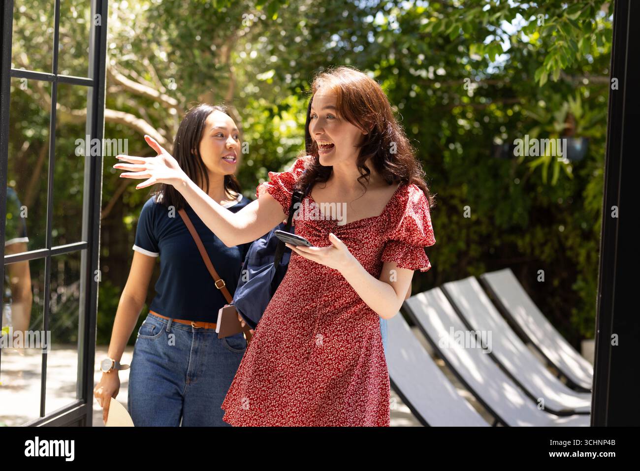 Zwei junge Frauen, eine mit langen braunen Haaren in rotem Kleid, betreten glücklich die sonnendurchflutete Terrasse Stockfoto