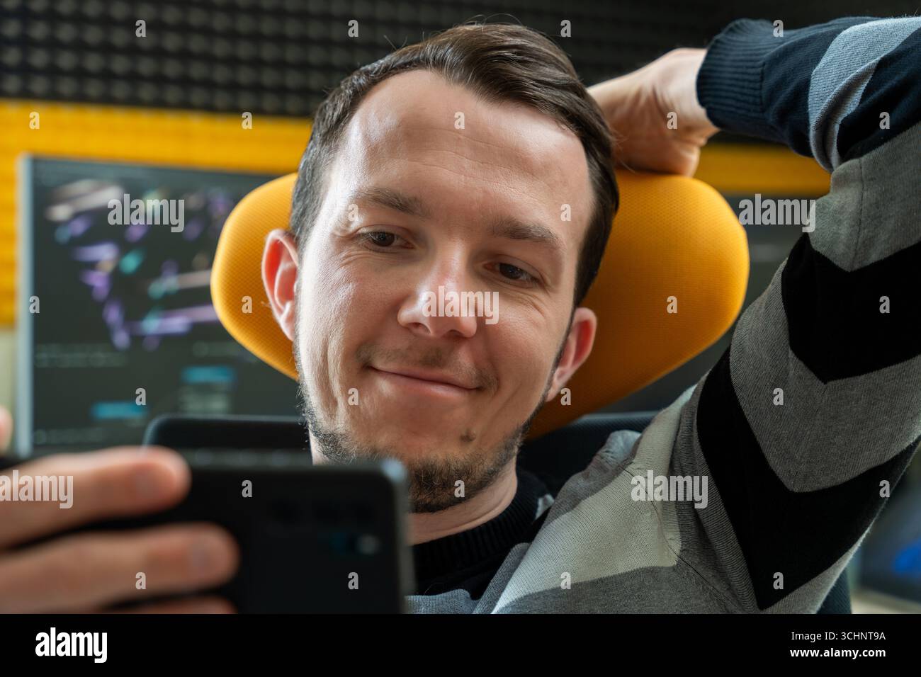 Lächelnder Mann, der sich in einem ergonomischen Stuhl zurücklehnt und im Büro mit Code auf dem Bildschirm auf das Smartphone blickt, Konzept der Zufriedenheit bei der Fernarbeit Stockfoto