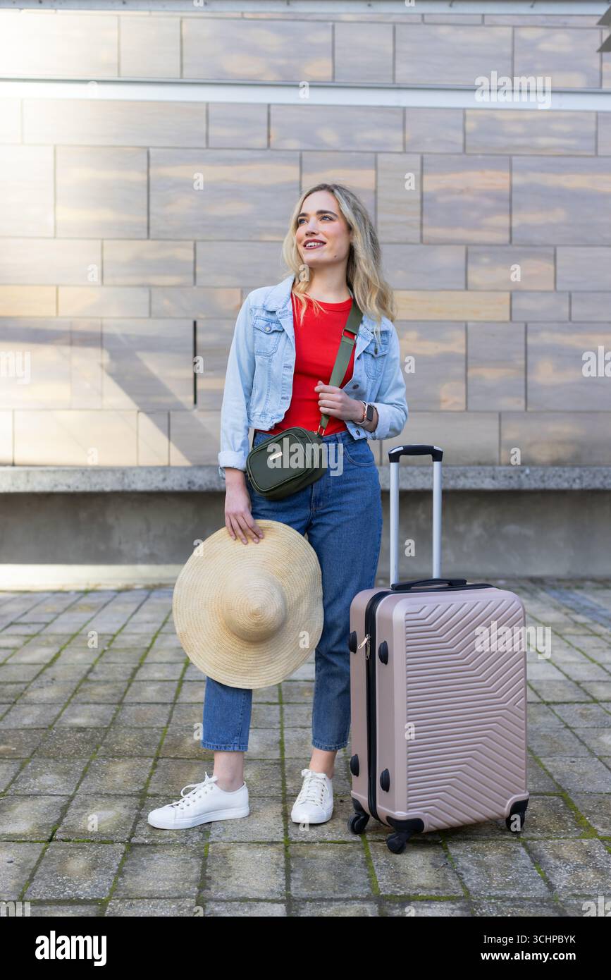 Frau zieht Rollkoffer und hält Strohhut auf der plaza mit grüner Tasche an die Steinmauer Stockfoto