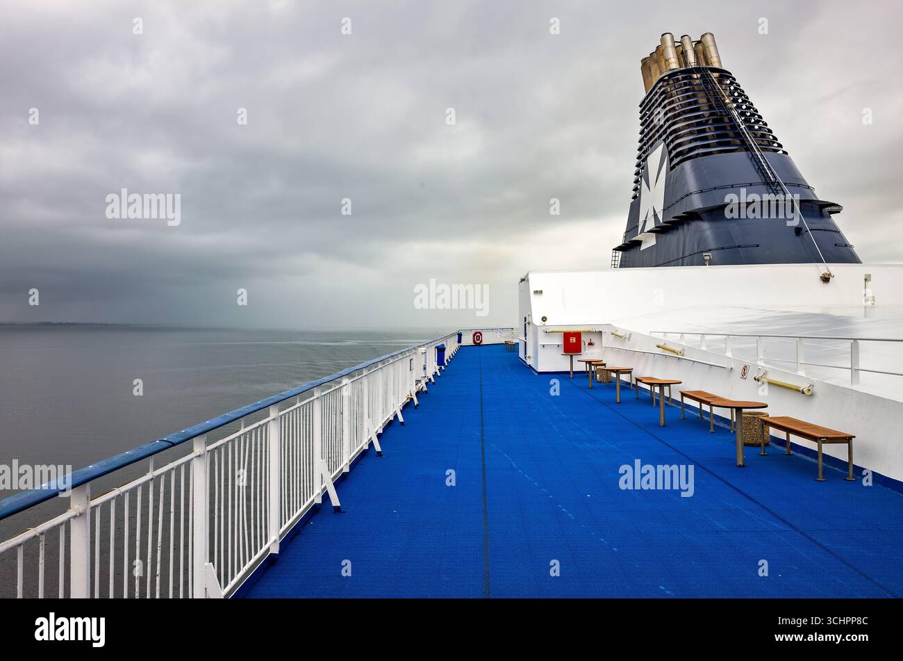 DFDS Ferry auf dem Oresund zwischen Rydebäck und Kopenhagen // RYDEBÄCK, Schweden — Eine DFDS-Fähre fährt über die Meerenge von Øresund zwischen Rydebäck, Skåne, Schweden und Kopenhagen, Dänemark. Das Deck der Fähre ist mit blauem rutschfestem Boden und weißen Geländern ausgestattet, mit Holzbänken und Tischen für Passagiere. Der markante Trichter des Schiffes, dunkelblau mit einem großen weißen „K“-Logo, erhebt sich vor einem bewölkten Himmel. DFDS ist ein dänisches integriertes Transport- und Logistikunternehmen. Stockfoto