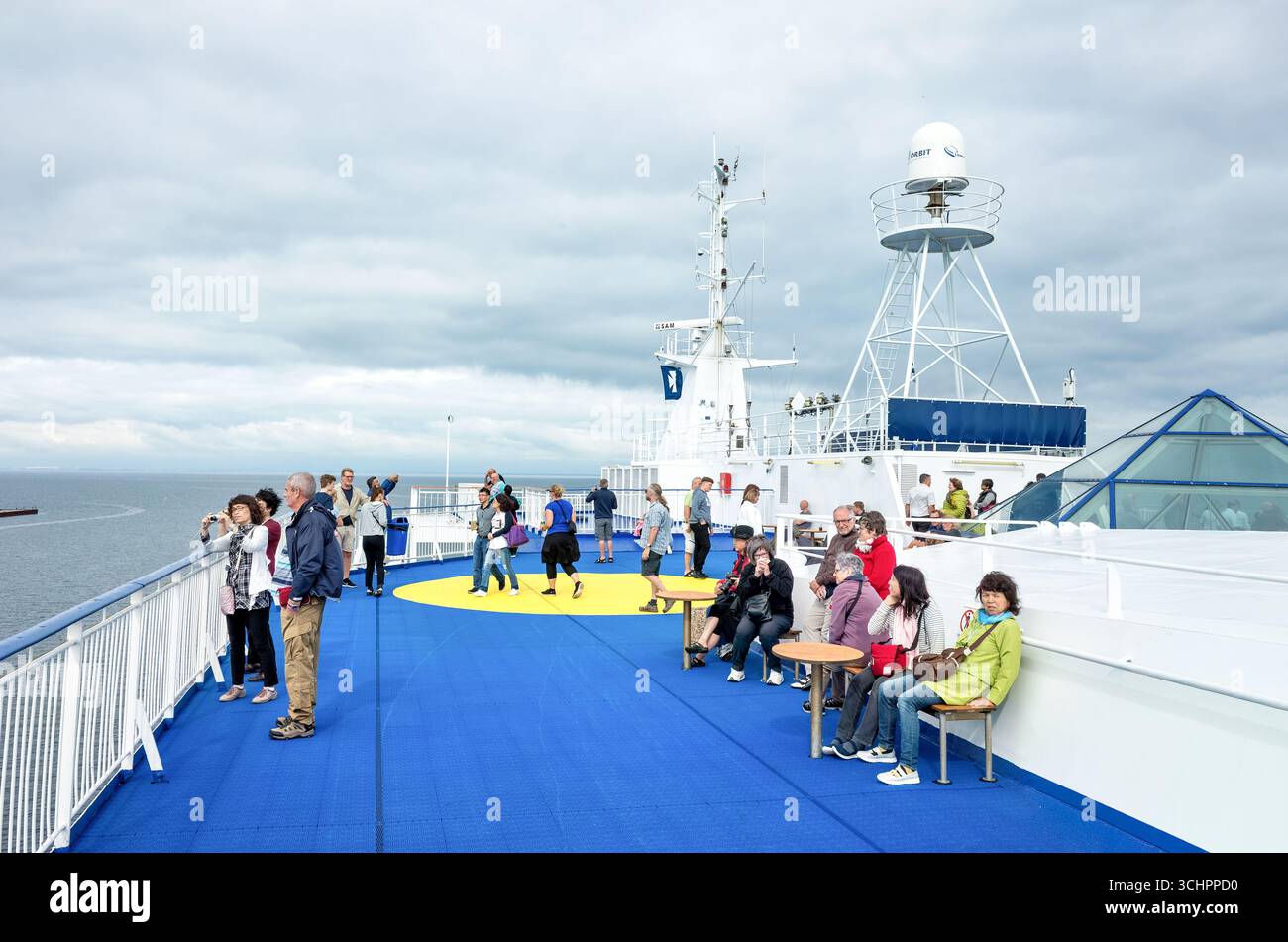 DFDS Ferry Passagiere auf Deck Øresund Dänemark // CHARLOTTENLUND, Dänemark — Passagiere entspannen sich auf dem offenen Deck einer DFDS-Fähre, während sie durch die Straße von Øresund fährt. Die Fähre verfügt über ein markantes blau-gelbes Deckmuster und ist mit verschiedenen Kommunikations- und Navigationsantennen ausgestattet, einschließlich einer auffälligen Radom-markierten UMLAUFBAHN. Die Øresund verbindet Dänemark und Schweden, und die Gewässer sind eine befahrene Schifffahrtsstraße. DFDS ist ein dänisches integriertes Schifffahrt- und Logistikunternehmen, das ein großes Netz von Fährstrecken und Logistikdienstleistungen betreibt. Stockfoto