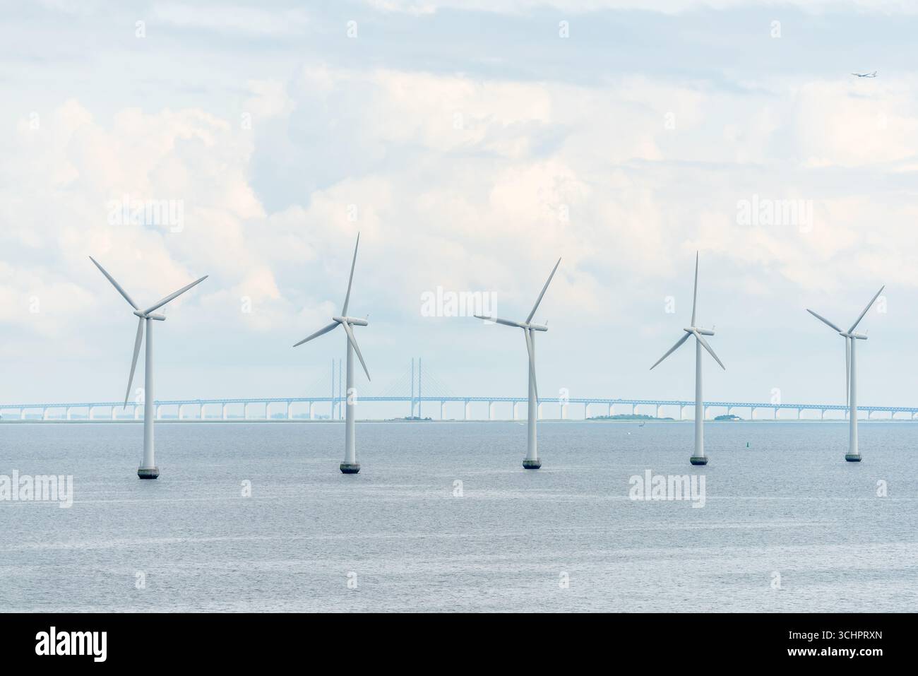 Windkraftanlagen Offshore mit Oresund-Brücke im Hintergrund Charlottenlund Dänemark // CHARLOTTENLUND, Dänemark — Offshore-Windräder sind in der Meerenge von Øresund sichtbar, im Hintergrund erstreckt sich die Øresund-Brücke über das Wasser. Die Brücke verbindet Dänemark und Schweden und überspannt die Meerenge zwischen der dänischen Insel Neuseeland und der schwedischen Provinz Scania. Dieses wichtige Infrastrukturprojekt ist eine bedeutende technische Leistung, die den Transport und den Handel zwischen den beiden Ländern erleichtert. Die Windturbinen stellen das Engagement Dänemarks für erneuerbare Energiequellen und Nachhaltigkeit dar Stockfoto