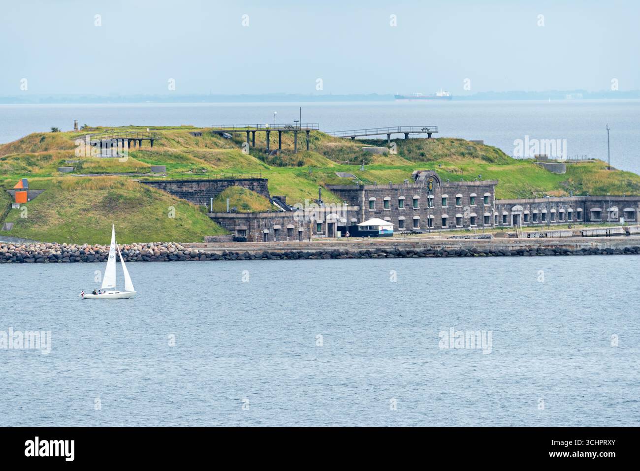 Flakfortet Charlottenlund Dänemark // CHARLOTTENLUND, Dänemark — Flakfortet, eine ehemalige Militärbefestigung, ist vom Wasser aus mit einem Segelboot in der Nähe zu sehen. Das Fort befindet sich in der Meerenge von Øresund nördlich von Kopenhagen und wurde zwischen 1910 und 1914 als Teil des dänischen Küstenverteidigungssystems errichtet. Es liegt auf einer künstlichen Insel und verfügt über eine markante Festung, die in die Landschaft eingebettet ist. Die Festung spielte eine Rolle in der dänischen Verteidigungsstrategie während der beiden Weltkriege. Heute ist Flakfortet eine historische Stätte und ein beliebtes Ziel für Freizeitaktivitäten. Das Kapital R Stockfoto