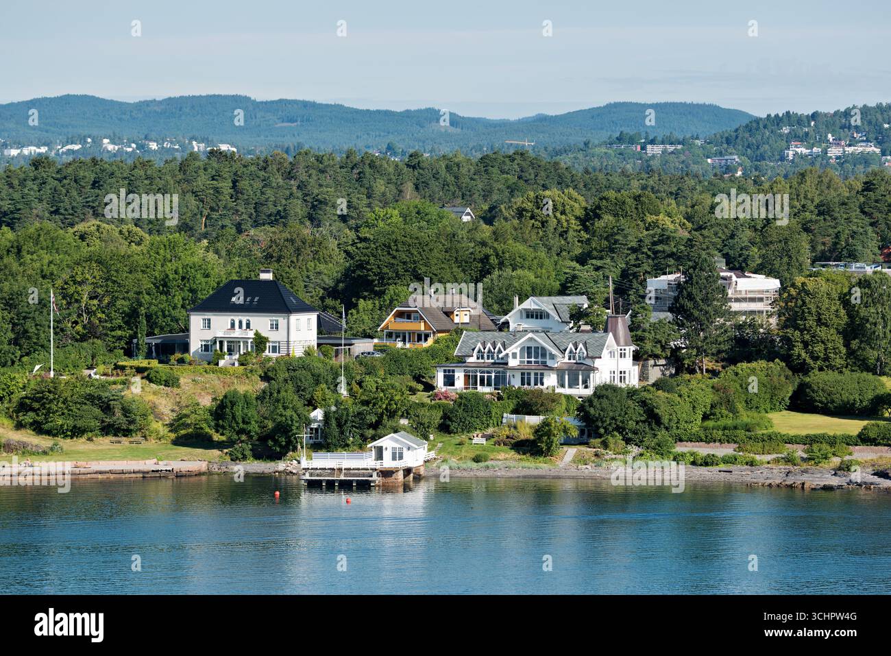 Häuser am Ufer des Oslofjords Lysaker Norwegen // LYSAKER, Norwegen — Häuser säumen die Uferpromenade des Oslofjords in Lysaker, Akershus. Die Wohngegend liegt eingebettet zwischen üppigen grünen Bäumen und sanften Hügeln. Mehrere große Häuser mit markanter Architektur sind entlang der Küste zu sehen. Der Oslofjord ist ein beliebtes Erholungsgebiet und eine wichtige Wasserstraße für die Region. Stockfoto