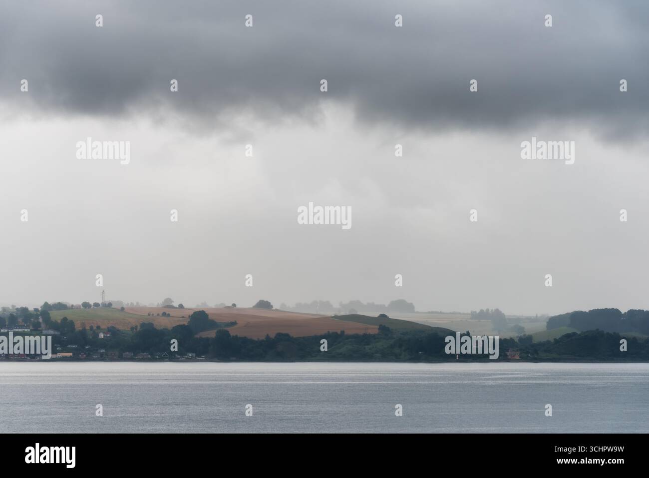 Küstenlandschaft mit Häusern und Hügeln Glumslöv Schweden // GLUMSLÖV, Schweden — Eine Küstenlandschaft mit sanften Hügeln und verstreuten Häusern entlang der Küste. Die Szene zeichnet sich durch einen Wasserkörper im Vordergrund und einen trüben, bewölkten Himmel aus. Glumslöv ist ein Dorf in der Gemeinde Landskrona in Skåne, Schweden. Die Region ist bekannt für ihre landwirtschaftliche Fläche und die malerische Küstenlandschaft. Stockfoto