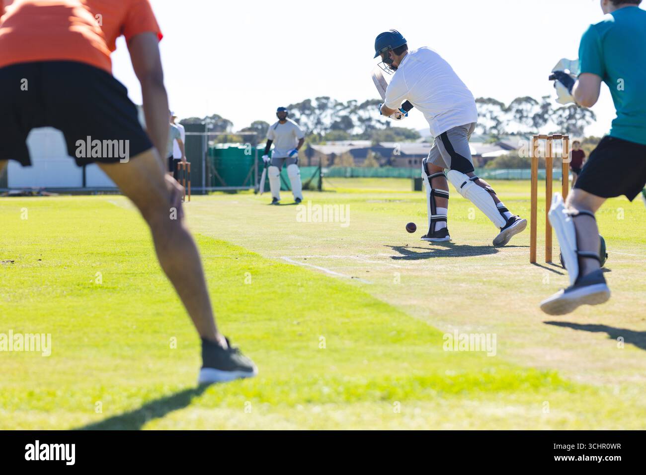 Männlicher Spieler schwingt Cricketschläger auf dem Grasfeld in Vorstadtgebieten, gegen Wicket, Feldspieler Stockfoto
