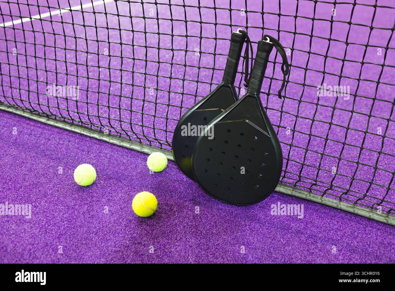 Zwei schwarze Padel-Schläger, die sich gegen das Netz auf violettem Rasen lehnen, mit gelben Kugeln, die verstreut liegen Stockfoto
