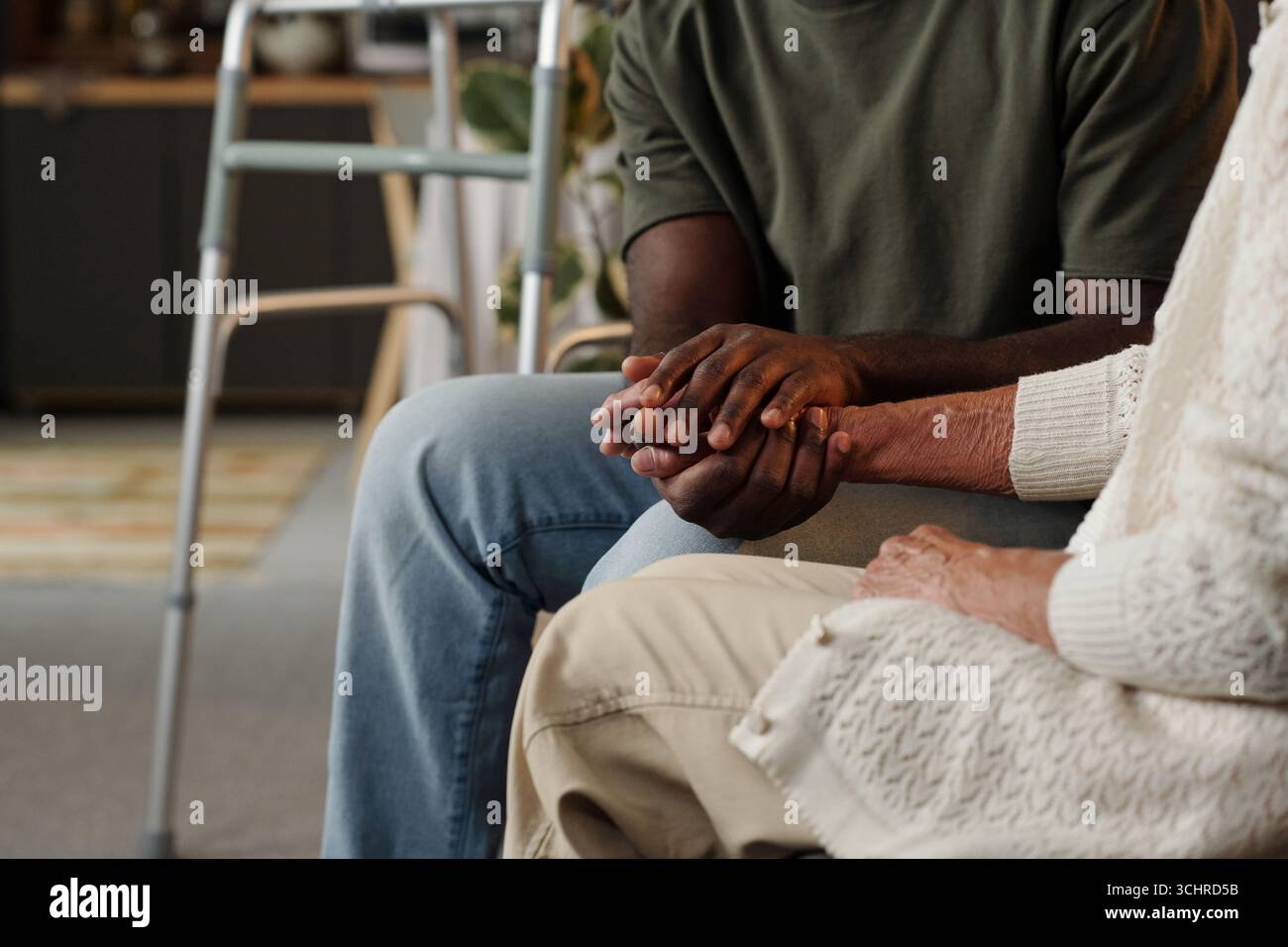 Schwarzer junger erwachsener Mann, der die Hand einer älteren kaukasischen Frau hält, die zusammen sitzt, zeigt Unterstützung und Mitgefühl, teilweiser Blick auf Hände und Arme, Wanderer sichtbar im Hintergrund Stockfoto