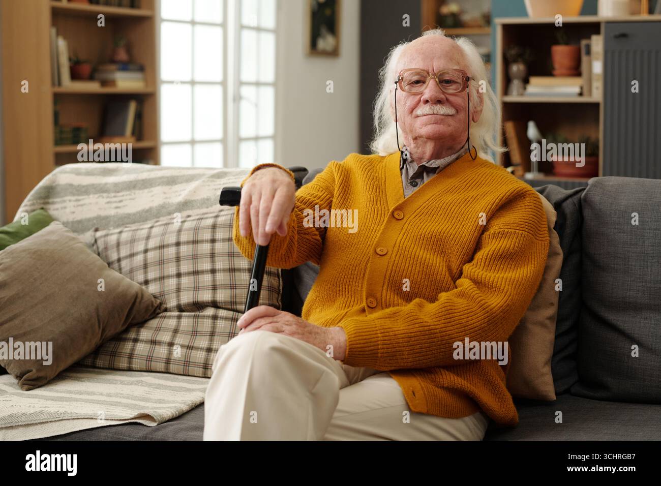 Porträt eines älteren kaukasischen Mannes, der auf dem Sofa sitzt und einen Gehstock mit der linken Hand hält, Brille trägt, mit ruhigem Ausdruck in die Kamera blickt, Haare und Schnurrbart sichtbar Stockfoto
