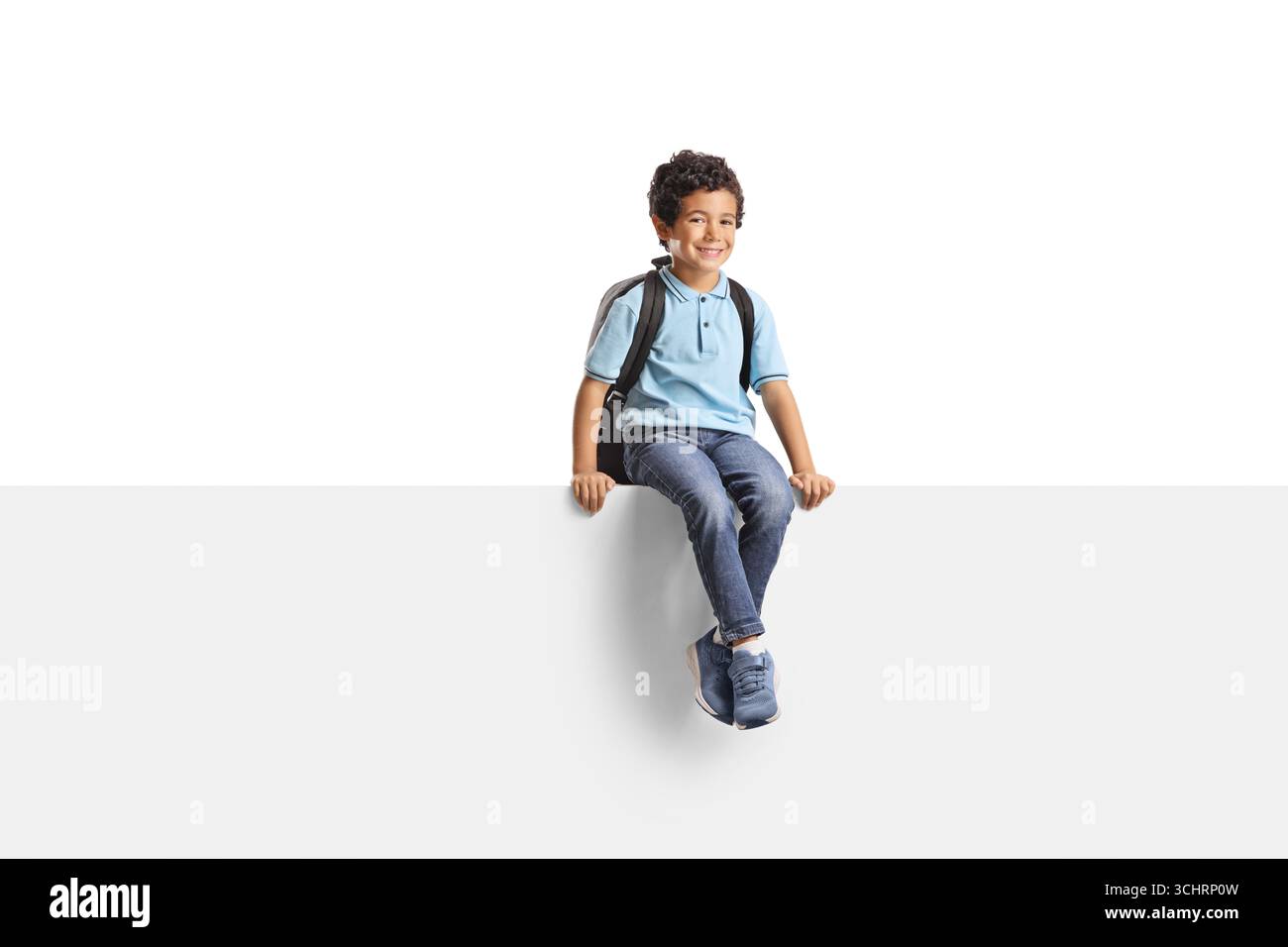 Junge mit einem Rucksack, der auf einer leeren weißen Tafel sitzt, isoliert auf weißem Hintergrund Stockfoto