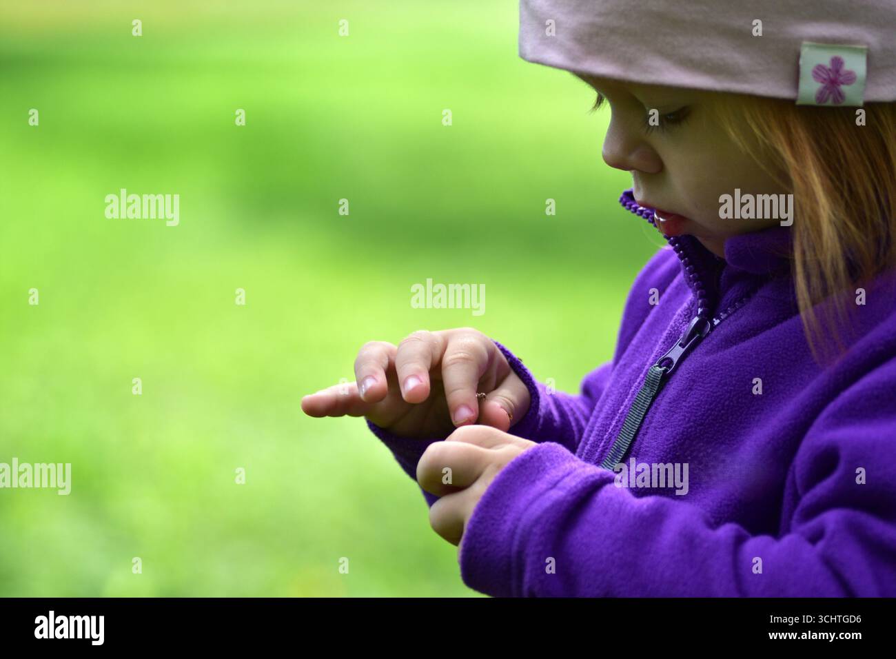 Nahaufnahme eines Kleinkindes in lila Jacke und rosa Hut, der ihre Finger im Freien sorgfältig betrachtet. Kindliches Detail und natürliche Neugier bei der Countryside Stockfoto