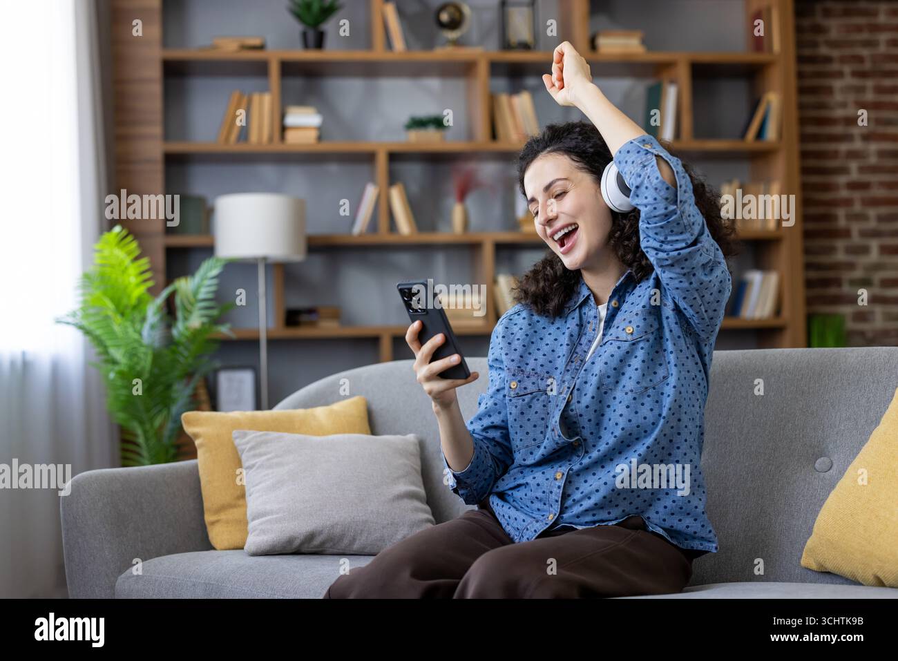 Junge Frau, die Kopfhörer trägt und Musik auf ihrem Smartphone hört, während sie auf einem bequemen Sofa in ihrem Wohnzimmer sitzt, ihre Lieblingslieder genießt und vor Freude tanzt Stockfoto