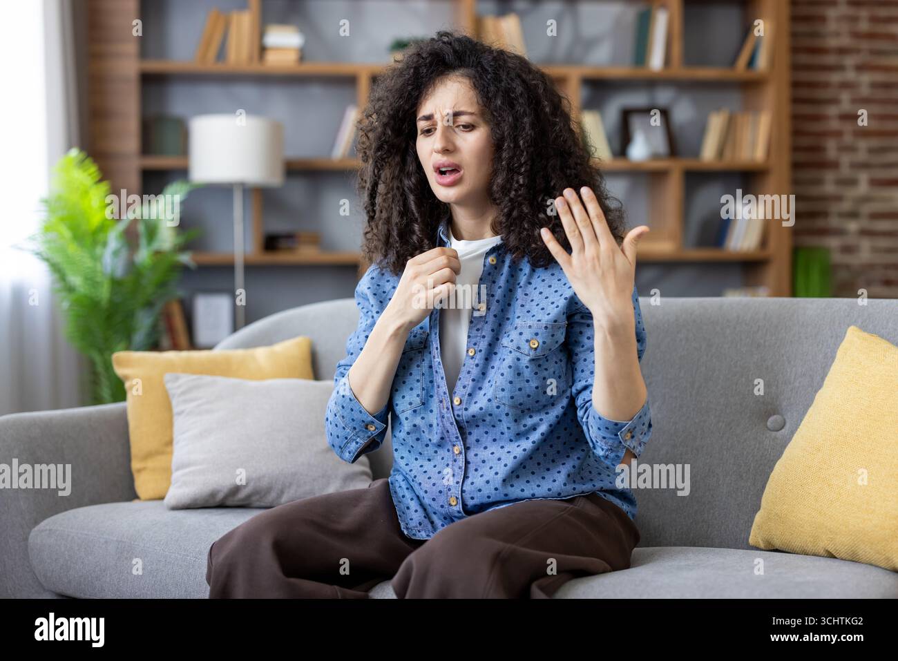 Junge Frau, die zu Hause auf dem Sofa sitzt und versucht, sich abzukühlen, leidet unter hohen Temperaturen und heißem Wetter, fühlt sich unwohl, fühlt sich unangenehm durch Hitzschlag im Sommer Stockfoto