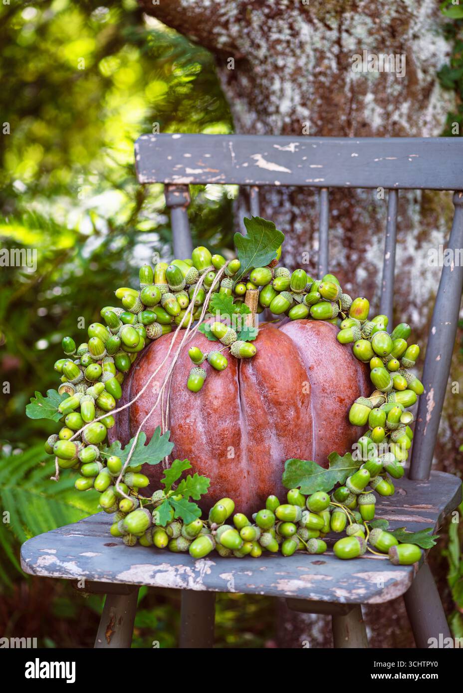Rustikales Stillleben im Herbst mit einem tieforangefarbenen Kürbis, gekrönt von einem handgefertigten, lebhaften Kranz aus grünen Eicheln, der auf einem verwitterten Stuhl unter einem Baum liegt. Stockfoto