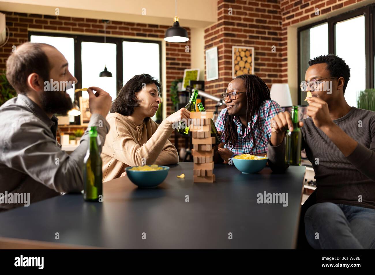 Multiethnische Gruppe von Freunden, die eine Pause von Spielen genießen, Snacks und Getränke am Tisch teilen. Kaukasische und afroamerikanische Frauen jubeln mit Bierflaschen, während Männer an Crisps knabbern. Stockfoto