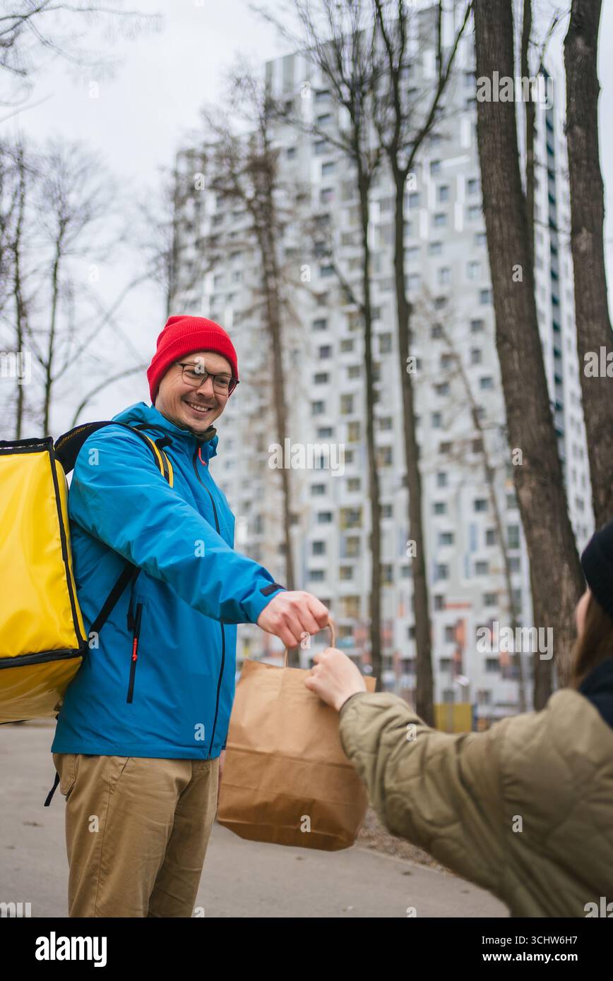 Mitarbeiter, der einen gelben Thermorucksack trägt, überreicht dem Empfänger eine Papiertüte mit Essen zum Mitnehmen in der Nähe von städtischen Grünflächen, was einen effizienten Service für die Lieferung von Lebensmitteln darstellt Stockfoto