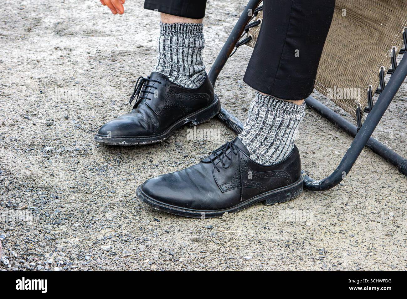 Ein Mann in formellen Schuhen und gemusterten Socken, der auf Kies steht und den Kontrast zwischen Formalität und Informalität im modernen Leben mit Selectiv darstellt Stockfoto