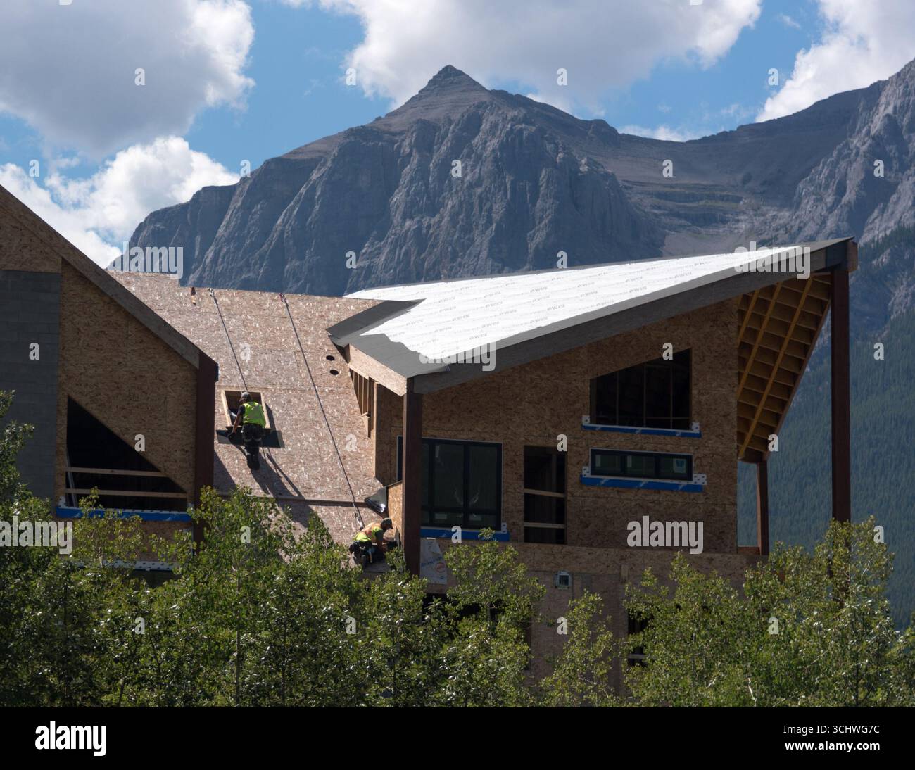 Zwei Bauarbeiter, die Kabelbäume verwenden, installieren Dachmaterialien in einem neuen Wohngebäude in Canmore, Alberta. Rundle Mountain bietet eine Kulisse. Stockfoto